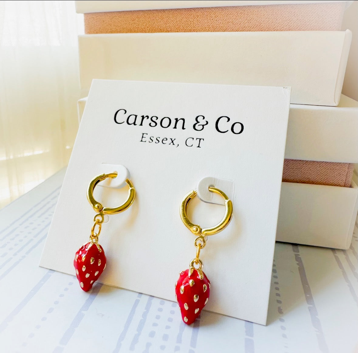A pair of Carson & Co strawberry drop earrings with gold hoop hoops and red strawberry charms, displayed on a white brand card.