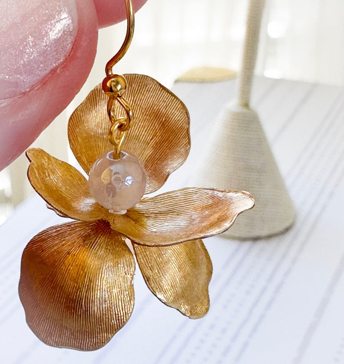 Gold flower-shaped earring with a pearl center held by a hand against a blurred background.