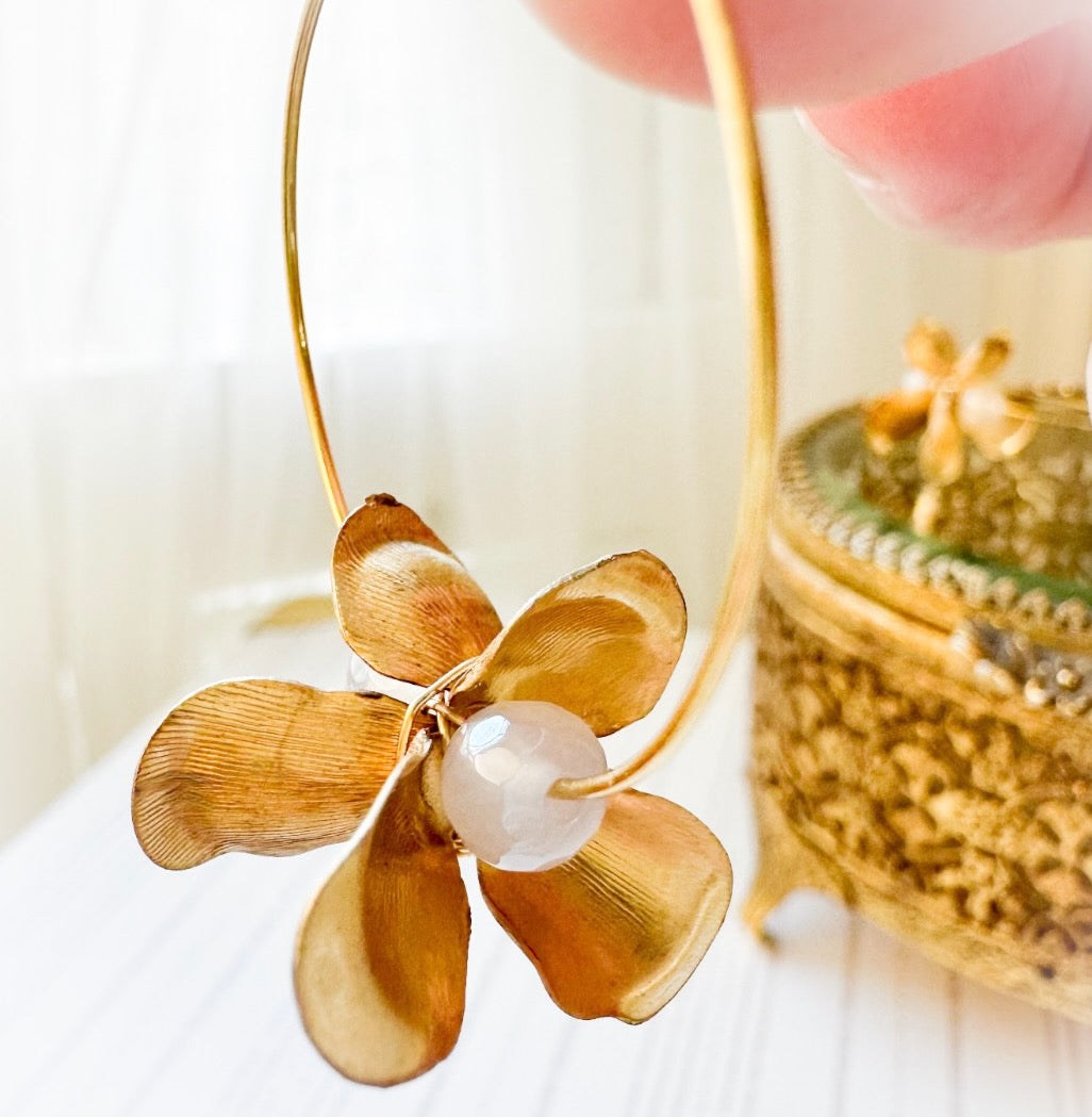Gold flower-shaped earring with a pearl center held by a hand against a blurred background.