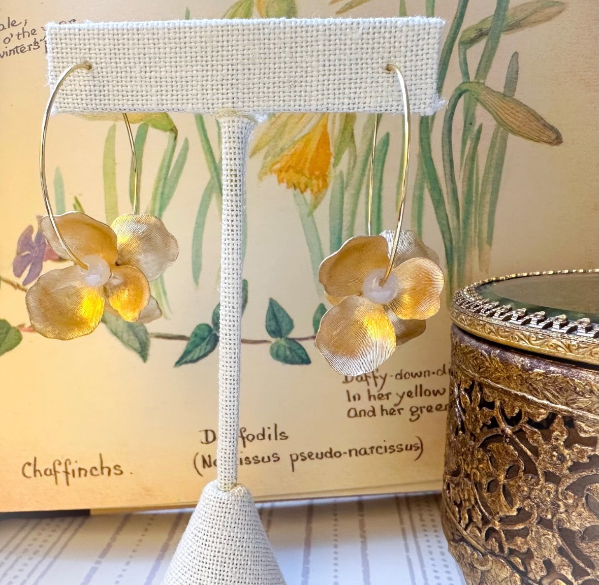 Gold hoop earrings with floral designs on a decorative stand against a nature-themed background.